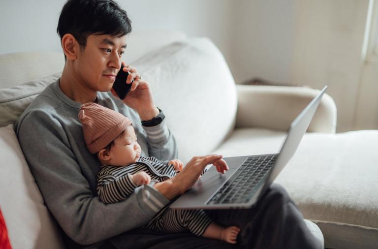 Father holding baby with laptop and phone.